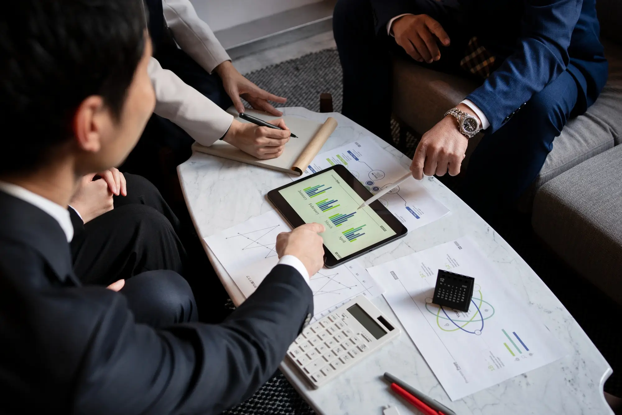 A table with an iPad, calculator, papers, and hands pointing at the items
