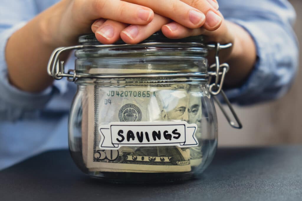 A jar labeled "savings" with cash inside and hands over the top of it