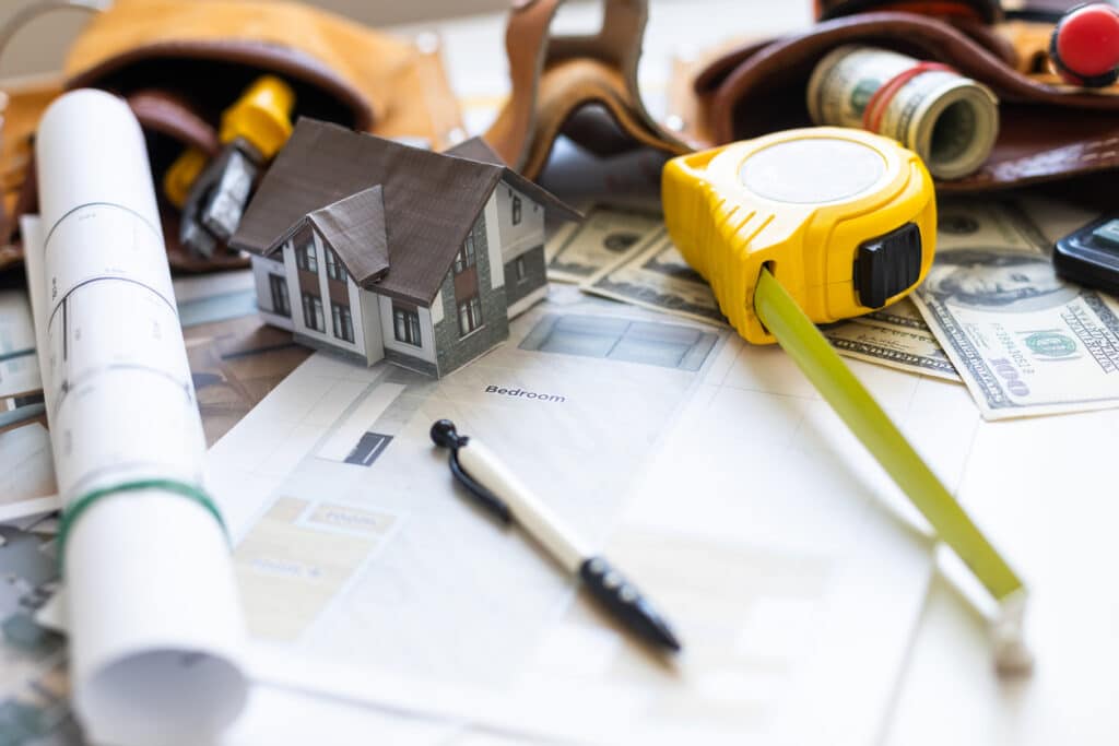 Table with cash, a measuring tape, a toolbelt, a blueprint, a model of a house, and a pen