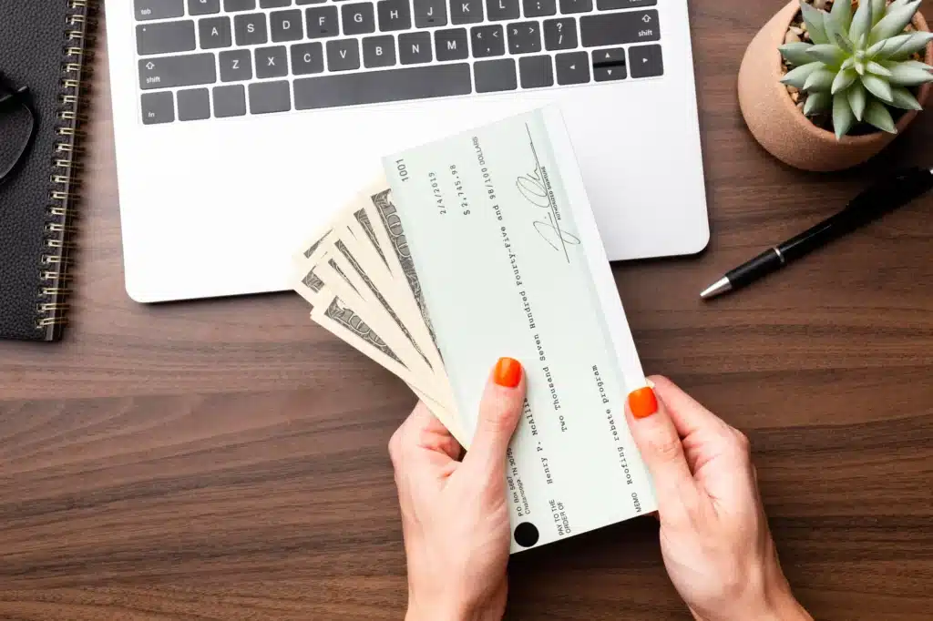 Hands holding cash and a check above a laptop